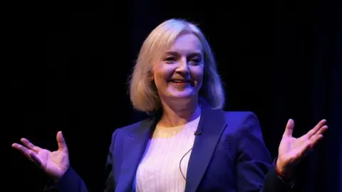 Liz Truss smiles and raised hands against a black background, on stage at Tory conference 