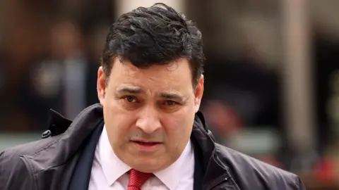EPA/Shutterstock A man with dark hair wearing a black jacket, navy suit, white shirt and red tie