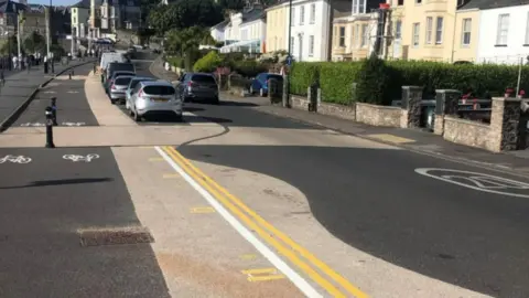 A picture of a road, with wiggly beige lines painted on the street and double yellow road markings. 