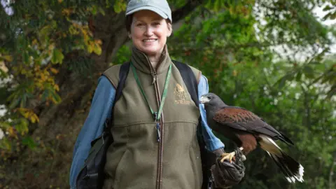 Facing the camera, Candida is slim with short brown hair, she is wearing khaki all-weather clothing, and has a whistle on a lanyard