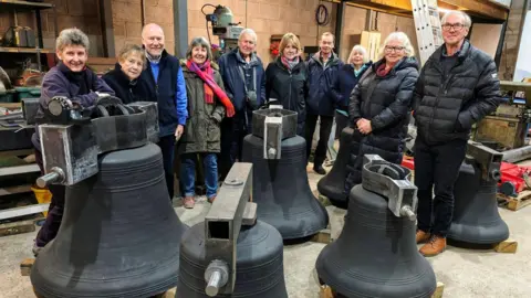 A group of 10 bell ringers standing in a warehouse building and smiling at the camera. They are gathered in a line, posing for a photo behind the restored bells, which are clean and black in new metal frames. 