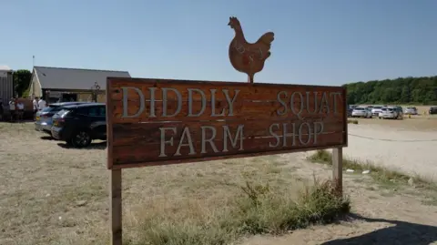 Getty Images A wide wooden sign with the words "Diddly Squat Farm Shop" spelled out in metal lettering. A wooden representation of a chicken in profile rises from the top of the sign. In the background are various parked cars and a building where several people are gathered. 