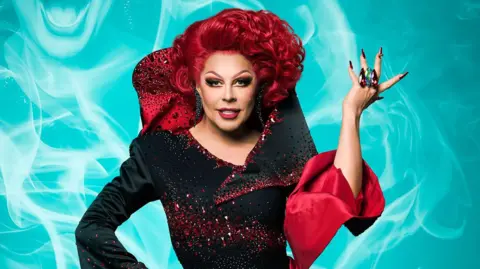 La Voix is wearing a black dress where the sleeves and collar resemble red emerging rose petals. Behind her is a digital blue background with white smoke.