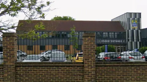 Google A school building with a fence and car park in front of it.