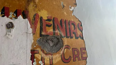 An orange sign with red lettering reading 'uvenirs' peaks through white paint, a large whole reveals the grey granite wall behind the sign. 