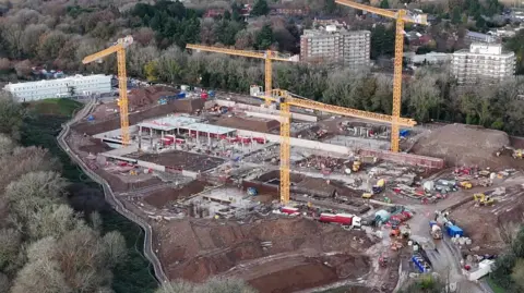 Construction work on the new hospital site filmed by a drone. There are four orange cranes with buildings in various stages of construction below them, some with roofs supported by pillars and others at the foundation stage. The site is surrounded by trees and there are two tower blocks of flats at the top right hand side of the picture.