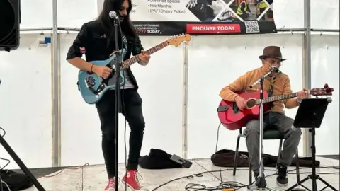 Kieran Bott Bansal Electrics are two people with one pictured standing playing a guitar and the other seated with a guitar. The man seated is wearing a brown jacket and grey jeans and the other band member is all in black with red trainers.