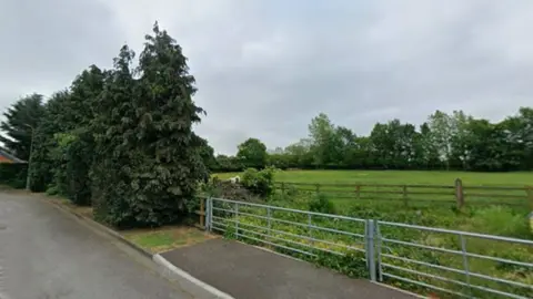 A paddock beyond wide metal gates and, behind those, a stretch of wooden fencing. The grass is a lush, bright green and there are darker green trees beyond. In the foreground is a tarmacked road and an edge of pavement. Trees line the road.