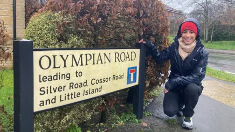 Shelley is stood next to a road sign displaying the name Olympian Road. She is wearing a red hat and blue raincoat.