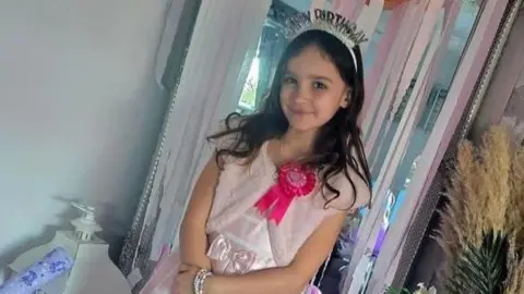Aria Thorpe is wearing a tiara which has "happy birthday" written on it. She is wearing a pink dress and has a pink lapel on.
