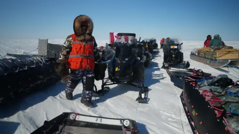 Eloise Alanna/BBC Barnie Aggark, in a grey and white camouflage snowsuit and fur hood, as well as a neon orange work vest, walks in the snow between sleds and snowmobiles. 