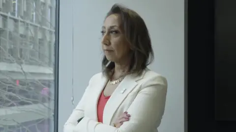 Moni Mannings, a woman with short brown hair wearing a cream jacket and coral top, looking out of the window of an office building with her arms folded