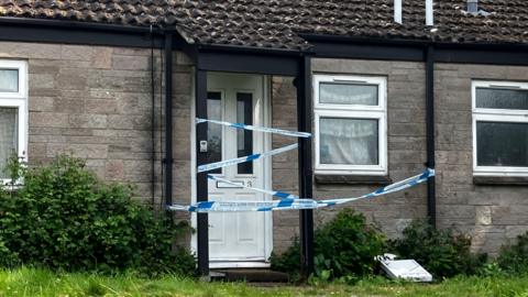 A picture of the outside of the home in Dunsford Terrace. The home appears to be a bungalow, with grey brick and blue and white police tape tied around the front door. 