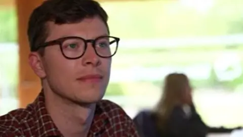Fred Best is wearing dark-rimmed glasses and has short brown hair and is wearing a red checked shirt. He is sitting down and looking off to the side and there is a woman blurred in the background, sitting at a desk.