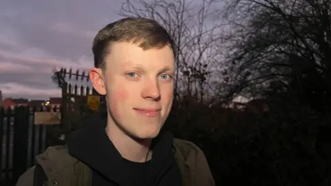 A young man smiles at the camera at twilight in Spark Mill Lane