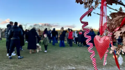 A crowd gathers on a field in the background, with a heart decoration hanging from a thin branch in the forefront 