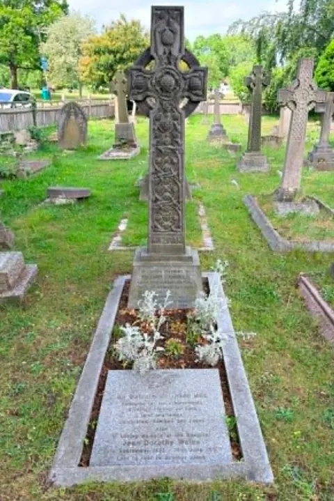 Steve Ladner A carved Celtic cross headstone in a grassy cemetery, with other older gravestones, trees, and a fence around it.
