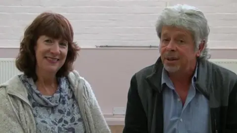 BBC A woman and a man smile at the camera. The woman has shoulde-rlength brwn hair and wears a floral jumper and grey zip-up. The man wears a blue shirt and grey and black zip-up jacket.