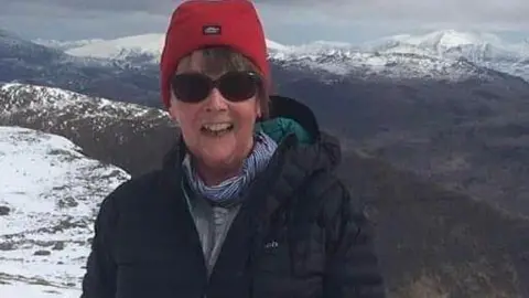 RoyCastle Sally Hall standing on top of a snowy mountainside wearing a red hat and dark sunglasses wearing a coat and scarf and smiling. 