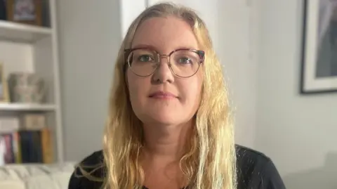BBC Emily Barley is sitting on a sofa in her lounge and she is holding a piece of paper which has two small handprints printed on it. Ms Barley has long blonde hair and is wearing glasses and a black top. There are framed photographs on the shelves to her right.
