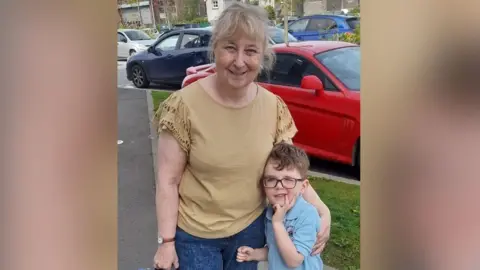 Darlene Garston who has light coloured hair tied back has her arm around her grandson Bertie who is wearing a polo top and has brown hair and glasses