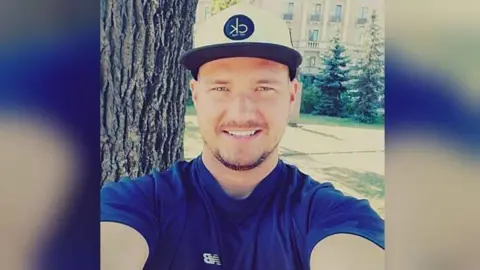 Police Scotland Oskars Rancevs smiles into the camera while standing outside on a sunny day. He is wearing a blue top and white baseball cap.