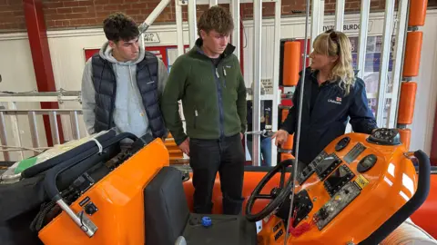 The brothers in the boat being shown how it works by RNLI volunteer Sarah Whitelaw. She has long blond hair and is wearing a navy RNLI jacket