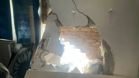 Chloe Beale The damage from the inside of the house. The white plaster has been cracked and damaged. Behind it is damaged brickwork and a hole through which daylight pours. A shelf has fallen to the floor, as has a grey painting of a flower. The patio door and window has a blue blind pulled down.