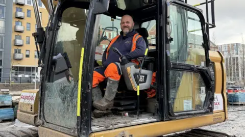 BBC / Becki Bowden The photo shows a man smiling at the camera. He's sitting in an orange digger cab, and is wearing a navy jumper and matching hi-vis jacket and trousers. The digger is in front of a block of flats.