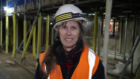 Shaun Whitmore/BBC Ruth Brennan, an architect, standing inside the St John the Baptist Church in Harleston. She is looking directly at the camera and smiling. She is wearing a zip up jacket with an orange fluorescent hi-vis vest over the top and a white hard hat.
