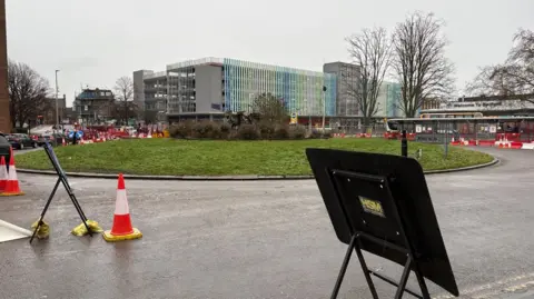 Martin Heath/BBC Green grass roundabout with trees in the centre - the sculpture is just visible behind them. In the foreground, there is a road on which a temporary roadsign has been erected. Cones and further signs are also visible.