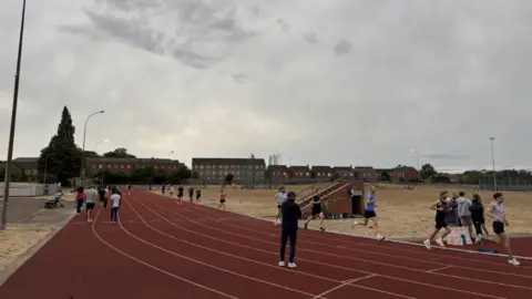BBC / Scott Dalton The photo shows a brown running track. There are a number of athletes running on it. There are a row of houses in the distance, as well some grass in the middle of the track. 