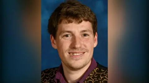 Sam Rabey smiles at the camera, he had brown hair and is wearing a black waistcoat with gold decoration.