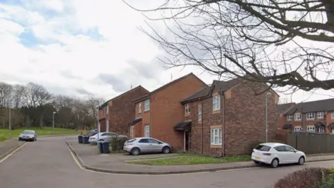Several brown and darker brown-brick houses can be seen in rows, some with cars parked outside them. A grass verge can be seen in front of them. 