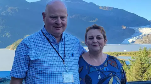 Nigel Walker Nigel and Penny Walker pose together on a trip with a mountain and lake behind them in the sunshine. He wears a blue check shirt and she wears a darker blue top.