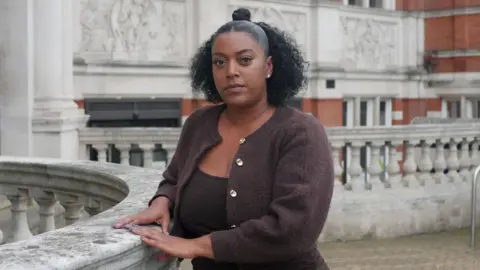 Nadine is standing outside, looking at the camera. She has black curly hair and is wearing a brown cardigan.