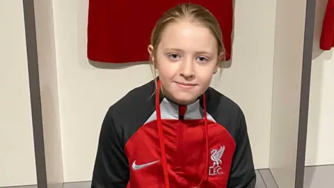 Girl with blonde hair wearing a red and black Liverpool Football Club jacket. She has blue eyes. 