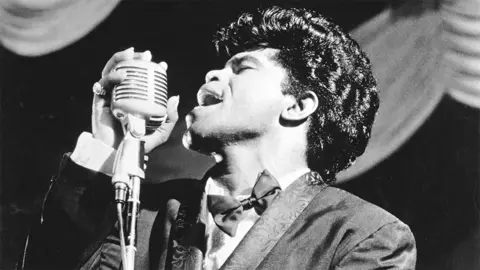 Getty Images James Brown bernyanyi ke mikrofon vintage saat ia tampil di atas panggung pada tahun 1962 di New York, mengenakan jas dan dasi kupu -kupu.