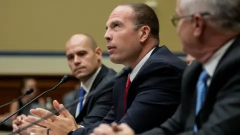 Getty Images Three men, all wearing dark suits and are seated at microphones.