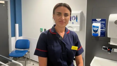 A photo of a woman with a blue short sleeved collared shirt on with red lining on it. She has brown hair tied up and has a little smile.