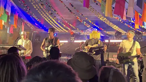 The Silver Bars The band on a small stage which is decorated with colourful bunting. On the left, one of the members is behind a keyboard and wearing a cream bucket hat. Next to him on the right is a man with brown hair who is wearing a vest top and is playing a guitar. Next to him is another man playing a bass guitar who is wearing a yellow hat with fringe around the brim. On the right is a man playing a guitar and wearing a green bandanna around his head. 