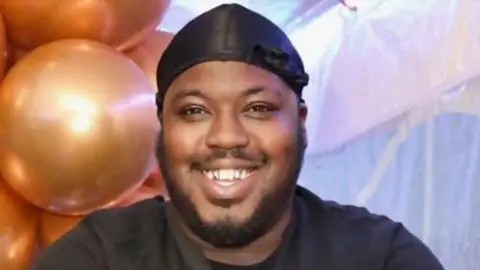 Family A man with a beard is smiling at the camera. He is wearing a dark top.