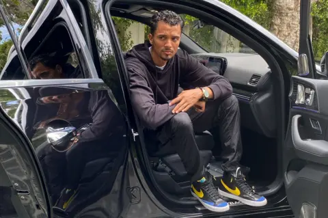 Phil Pines in a black hoodie, pants, and sneakers with yellow and blue accents sitting in the passenger seat of a black car with the door open.