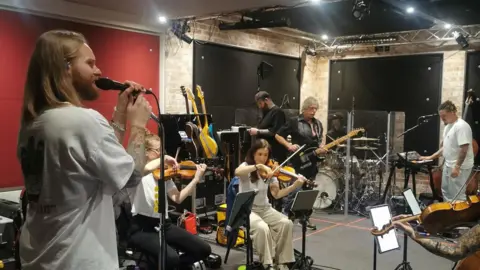 Kelvin Pratt Sam Ryder singing with his live band. People are playing a violin while sitting down. While other people are playing the guitar. Sam is on the left of the picture and holding a microphone. They seem to be in a studio rehearsing.
