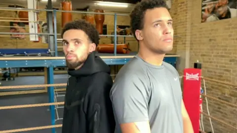 Two men in a boxing gym standing back to back, with a boxing ring behind them. 