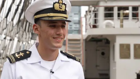 A smiling young naval cadet wears a smart white shirt with black epaulettes on the shoulders that have gold anchors embroidered on them. He wears a white military cap with black peak and gold embellishment. The bridge of the ship is behind him in the background