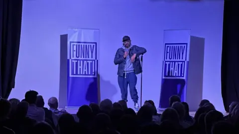 Deeps Motley Ranganathan performs at the village hall. The stage is lit up in a purple blue light and there are signs for the production company Funny That, which staged the event. People in the audience are sitting in the dark.