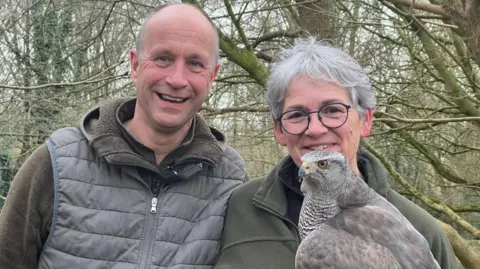 A couple wearing khaki green and brown clothing in woodland in winter. On the left, a man with grey hair smiles at the camera, alongside his wife to the right, who also has grey hair and navy glasses. She is holding a mottled brown, grey and white Goshawk which looks to the left beyond the camera. It has piercing orange and black eyes and a hooked beak. 