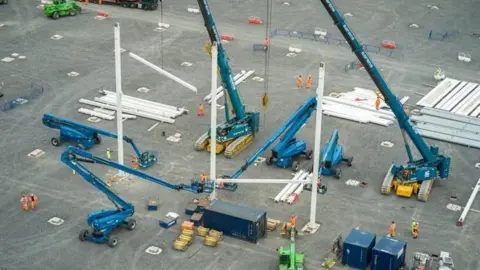 A series of blue cranes on a construction site lifting up a steel structure with construction workers in orange high-vis clothes on the ground.