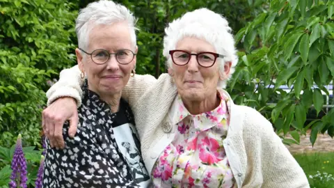 Jean Finch Anita and Steph, standing outside in a garden, with plants and trees. They are both smiling and looking straight at the camera. They both have glasses. Anita is wearing and black and white top, Steph, a floral top with a cream cardigan. 